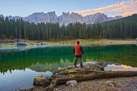 A man stands on a stone by the lake, watching the sunrise over the mountainsの写真素材
