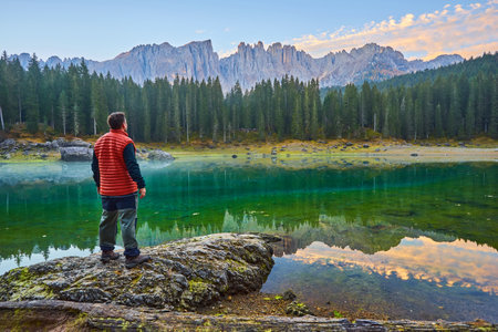 A man stands on a stone by the lake, watching the sunrise over the mountains