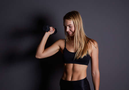 Blonde athletic woman shows her trained body on a dark background. female model trains triceps with dumbbells. healthy lifestyle and sport concept.の写真素材