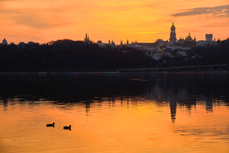 Kiev-Pechersk Lavra and city lights, in the evening. Silhouette panorama at sunset of the colorful sky over the Dnieper river with passing ducks in Kyivの写真素材