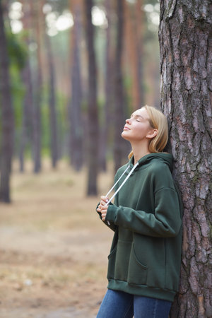 Cheerful blue-eyed blonde girl in green hoodie, smiles against park background. Attractive blonde in stylish clothes posing outdoor. Relax in nature.の写真素材