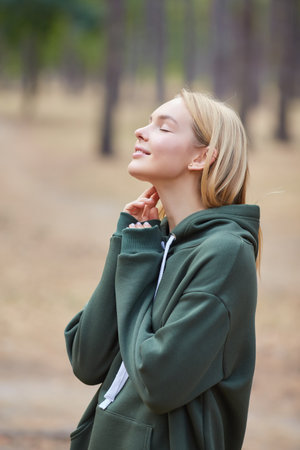 Attractive blue eyed blonde woman walk on the city park. Girl wear green hoodie, look happy and smiley. Portrait of a joyful young woman enjoying in autumn park.の写真素材