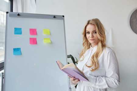 A blonde girl stands near a board with stickers and holds a document and a pencil in her hands....