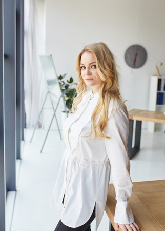 professional and beautiful. Cheerful young woman stands by the window leaning on a wooden table and looking at camera with smile while standing at her working placeの写真素材