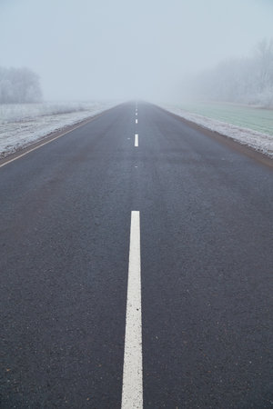 Winter morning with frosted trees, foggy atmosphere, and a disappearing asphalt road. An asphalt road stretches into the distance, disappearing into the mist, adding to the enigmatic composition.の写真素材