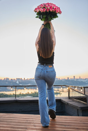 Young brunette with a large bouquet of roses takes a photo on the rooftop, with the city, river, and...
