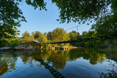 old small wooden plank bridge over river in countryside, sunny summer day, green trees in background