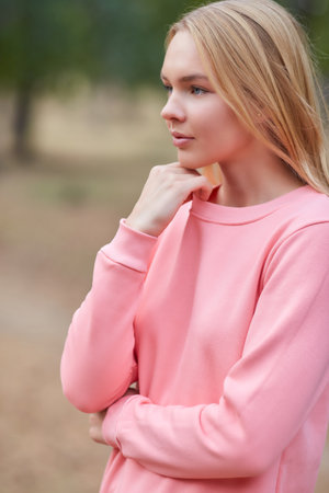 Blue-eyed blonde in a pink hoodie walks in a pine forest. Portrait of a joyful young woman enjoying in autumn park. Relax in nature.の写真素材