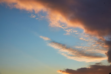 A calm sky with soft golden clouds illuminated by the setting sun. The subtle transition from bright blue to warm orange creates a peaceful and dreamy atmosphere over the horizon.の写真素材