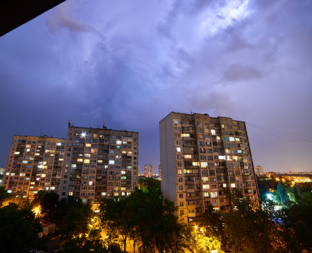 A nighttime cityscape illuminated by streetlights and building windows, with a dramatic streak of lightning cutting through the cloudy sky. The urban landscape contrasts with the stormy atmosphere.の写真素材