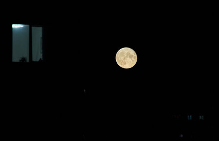 A bright full moon shines in the dark night sky, accompanied by the glowing window of a nearby building. The simplicity of the scene contrasts the natural moonlight with artificial indoor light.の写真素材