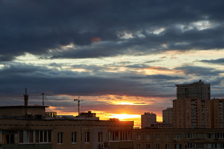 A moody sunset with clouds rolling over a city skyline. The sun peeks through the clouds, casting a warm glow over the buildings, contrasting with the darkening evening sky.の写真素材