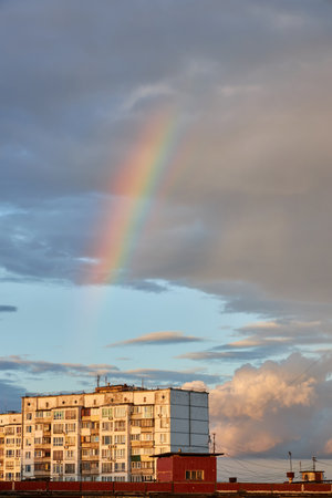 A vibrant rainbow appears in the sky above a city building after a rainstorm. The soft light illuminates the clouds and the building, creating a peaceful post-storm urban scene.の写真素材