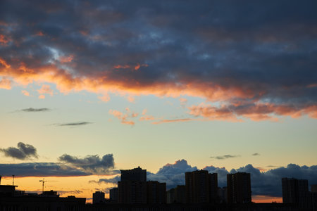 A moody sunset with clouds rolling over a city skyline. The sun peeks through the clouds, casting a warm glow over the buildings, contrasting with the darkening evening sky.の写真素材