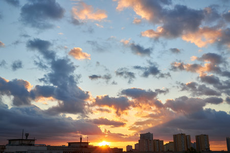A soft pastel sunset with light pink clouds floating above silhouetted rooftops and distant city buildings. The warm evening sky transitions from yellow to light blue, creating a peaceful urban scene.の写真素材