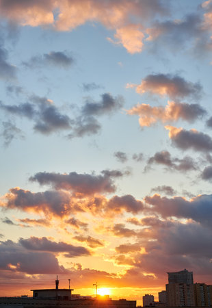 A vibrant sunset with intense orange and yellow hues highlights the silhouettes of buildings and a construction crane, creating a dramatic skyline under a cloudy sky.の写真素材
