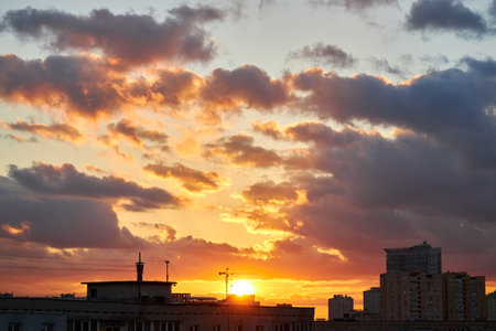 A moody sunset with clouds rolling over a city skyline. The sun peeks through the clouds, casting a warm glow over the buildings, contrasting with the darkening evening sky.の写真素材