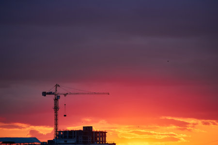 A construction crane silhouetted against a vibrant red and orange sunset sky. The glowing horizon creates a striking contrast with the dark urban elements, capturing the beauty of an evening cityscape.の写真素材