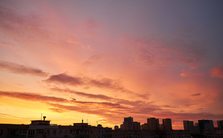A soft pastel sunset with light pink clouds floating above silhouetted rooftops and distant city buildings. The warm evening sky transitions from yellow to light blue, creating a peaceful urban scene.の写真素材