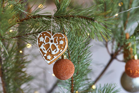 Close-up of a heart-shaped gingerbread cookie with white icing and a glittery orange bauble hanging on a pine branch. Warm festive lights add to the cozy holiday atmosphere.の写真素材