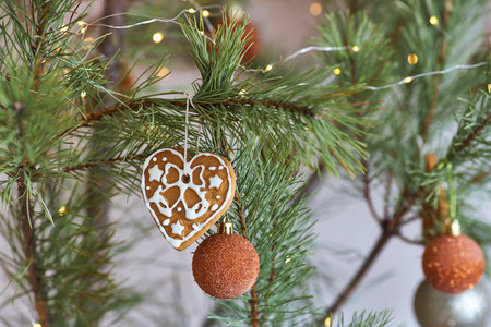 Close-up of a heart-shaped gingerbread cookie with white icing and a glittery orange bauble hanging on a pine branch. Warm festive lights add to the cozy holiday atmosphere.の写真素材