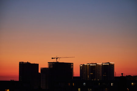 A city skyline at dusk with a gradient sky transitioning from warm orange to deep blue. The silhouettes of buildings and cranes are visible against the glowing horizon.の写真素材