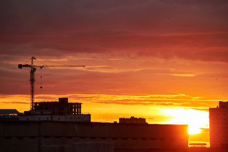 A bright golden sunset casts a warm glow over silhouetted buildings and a construction crane. The intense light filters through clouds, creating a dramatic and radiant urban sceneの写真素材