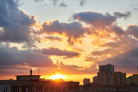 A vibrant sunset with intense orange and yellow hues highlights the silhouettes of buildings and a construction crane, creating a dramatic skyline under a cloudy sky.の写真素材