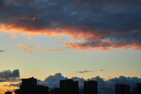 A vibrant sunset sky with dark clouds and orange highlights contrasts against the city skyline. The moody atmosphere captures the beauty of a changing sky over an urban landscape.の写真素材