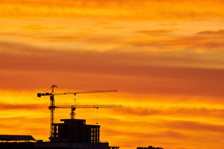 A bright golden sunset casts a warm glow over silhouetted buildings and a construction crane. The intense light filters through clouds, creating a dramatic and radiant urban sceneの写真素材
