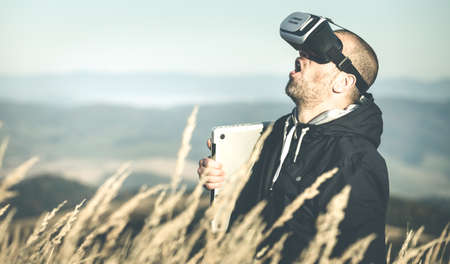Young man screams and looks upwards in virtual VR glasses - laptop under his arm and virtual goggles on his head as a new technology trend - millenials walks in the meadow with a virtual reality kitの写真素材