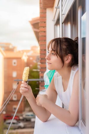 Young girl with face mask leaning out of the window eating ice cream during quarantine. Person confined in summer eating ice creamの写真素材