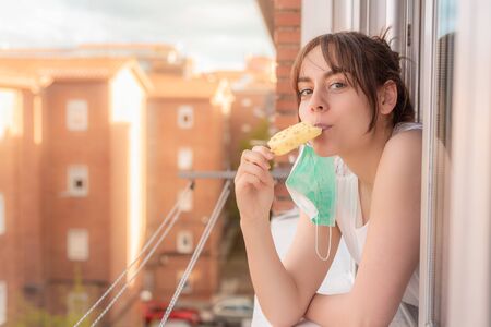 Young girl with face mask leaning out of the window eating ice cream during quarantine. Person confined in summer eating ice creamの写真素材
