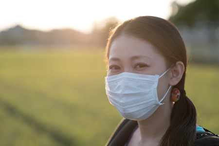 A young Japanese woman wearing a surgical mask outdoors in the countryside with a bright sunset behind her.の写真素材