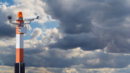 Two CCTV security cameras on red striped pillar with orange flashing light against the cloudy sky の写真素材