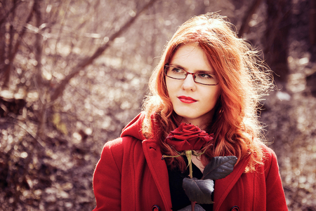 Outdoor fashion closeup portrait of pretty young ginger woman in red coat with rose; toned imageの写真素材