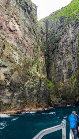 Super spectacular Vestmanna steep cliffs in the Faroe Islands with unrecognizable tourists over the boatの写真素材