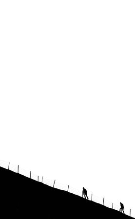 Two men silhouette hiking over high slope hillside under white sky with text spaceの写真素材