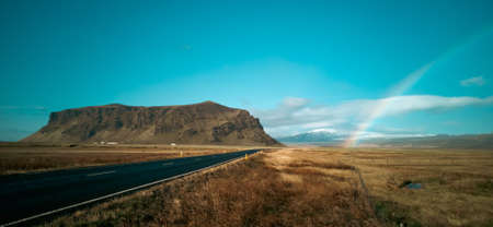 Straight road, rainbow and mountains under clear blue skyの写真素材
