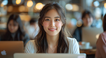 Young Asian woman smiling at camera in a busy cafe with laptop in front of her.の素材