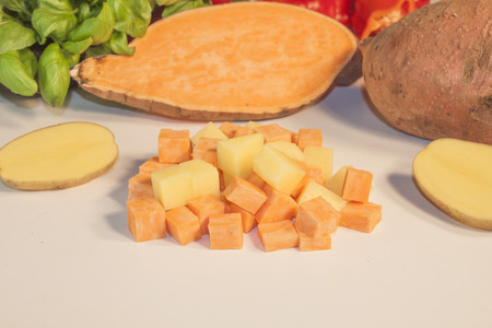 Sweet potato and potato dice, in front of herbs and vegetablesの写真素材