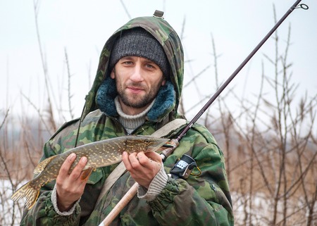 Happy fisher, dressed in old camouflage jacket, with catched by spinning pike in his hands, late autumnの写真素材