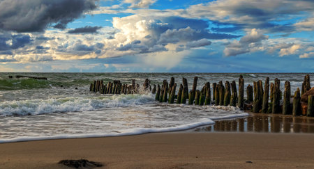Waves gently lapping against wooden posts on a serene sandy beach under a vibrant sky near the coastlineの写真素材