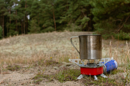 Portable camping stove with metal mug on a red base during daylight in a forest clearing surrounded...