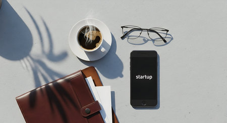 A clean and modern flat lay of essential work items: a steaming cup of coffee, stylish eyeglasses, a smartphone displaying 'startup', and a classic brown notebook, all arranged on a light gray backgr.の素材