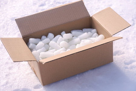 A brown corrugated cardboard box sits open in deep white snow, filled with numerous translucent white ice cubes. Bright daylight illuminates the frozen texture.