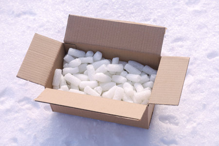 An open brown cardboard box sits on a snowy ground, filled with numerous white ice cubes. The scene is captured in bright daylight with sharp details.