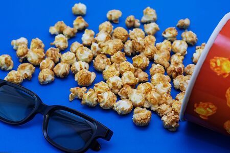Caramel popcorn scattered on a blue background and 3D glasses for watching a movie. The concept of the film. Food for watching movies. View from above. 2020 blue color background.の写真素材