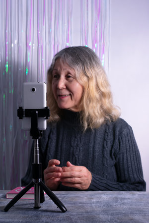 An elderly woman sits at a table in front of a smartphone on a tripod. Senior woman blogger is recording a video that is broadcast online indoorsの写真素材