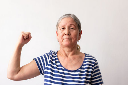 Elderly woman, 60s gray-haired lady in striped white and blue dress, strong man showing arm muscles, confident and proud of his power on white background.の写真素材